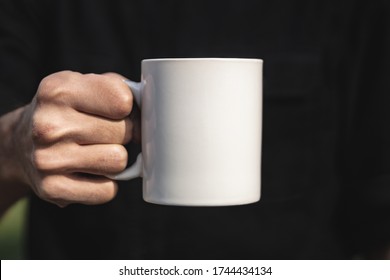 Man Holding A White Cup Of Hot Beverage Outdoors. Man's Hand Holding A Blank Mug Against Black Background. Close Up, Mockup Image, Copy Space For Text.