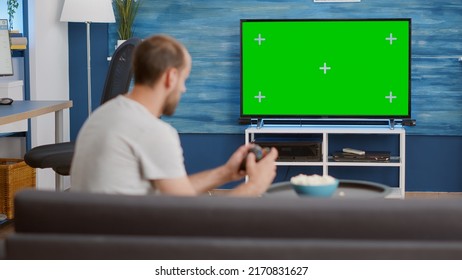 Man Holding Wireless Controller Playing Console Video Game On Green Screen Tv While Sitting On Sofa In Modern Living Room. Gamer Relaxing On Couch Enjoying Online Gaming On Croma Key Display.