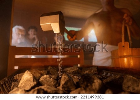 man holding wooden bucket and pouring water onto hot stones with rain spoon in sauna room with a group of people. Steam an water on the stones, spa and wellness concept, relax in hot finnish sauna.
