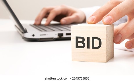 man holding wooden cube witt letters BID in the background a laptop on a white office table background