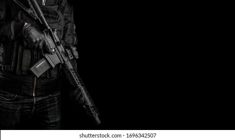 A man holds an automatic carbine with a collimator sight on a dark back. A fighter in dark clothing and a tactical vest with a weapon in his hands. Poster concept for police, security or military
