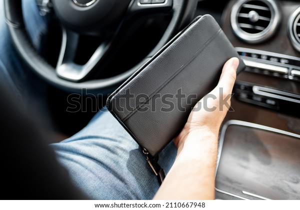 A man holds a black\
leather wallet in his hands while sitting on the driver\'s seat of a\
luxury car. A young businessman driving a luxury car with a wallet\
in his hand.