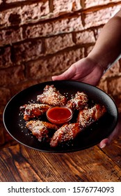 Man Holds Black Round Plate With Beautiful Fried Chicken Wings Sprinkled With White Sesame Seeds With Red Barbecue Sauce On Dark Brick Background. Close-up.