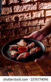 Man Holds Black Round Plate With Beautiful Fried Chicken Wings Sprinkled With White Sesame Seeds With Red Barbecue Sauce On Dark Brick Background. Close-up.