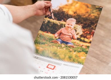 Man Holds A Calendar With A Photo Of A Child. Printed Products With An Image. A Gift To Parents And Grandparents From Children And Grandchildren For New Year. Services Of Photographer.