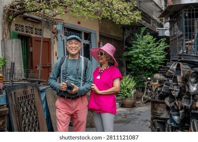 A man holds a camera while standing next to a woman in colorful clothing. They smile as they walk through a narrow street filled with plants and old items.