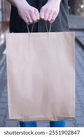 A man holds a craft paper bag by the handles while standing on the street