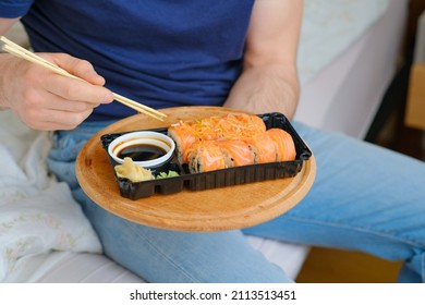 A Man Holds Delivered Sushi With Chopsticks And Plastic Box With Salmon Roll In The Bedroom. Cozy Home. Eating In Bed. Meal. Traditional Asian Food. Takeout And Delivery. 