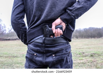 A man holds a gun tucked into his pants behind his waist in an open field.