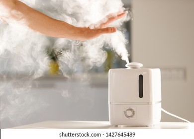 Man holds hand over steam aroma oil diffuser on the table at home, steam from the air humidifier, free space. Ultrasonic technology, increase in air humidity indoors, comfortable living conditions