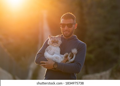 Man Holds In Hands His Lovely Purebred Cat Outside On The Sunset. Pet With Owner