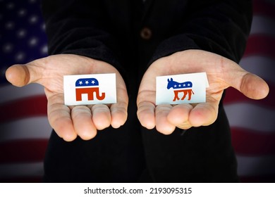 Man Holds In His Hands Elephant Symbol Of The Republican Party And The Donkey Symbol Of The Democratic Party, With The American Flag Behind. 13 August 2022