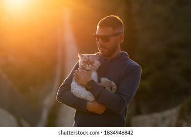 Man Holds His Lovely Groomed Purebred Cat Outside On The Sunset. Pet With Owner