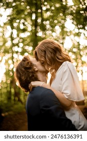 Man holds his wife in his arms and they kiss while standing against the background of the forest