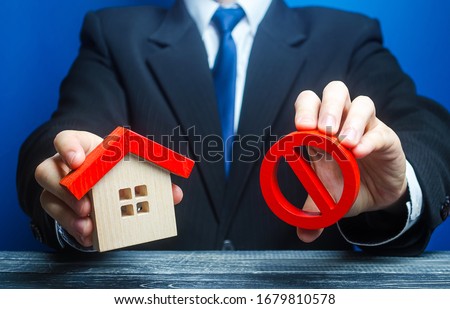 A man holds a house and the red prohibition symbol NO. Bank refuses to provide a mortgage loan. Housing problems. Confiscation pledged property. Prohibition of operation. Denial of insurance payment.