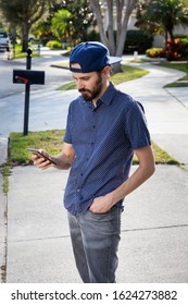 A Man Holds And Looks At Cell Phone, Standing At The Curb Of A Neighborhood Waiting For His Ride Share.