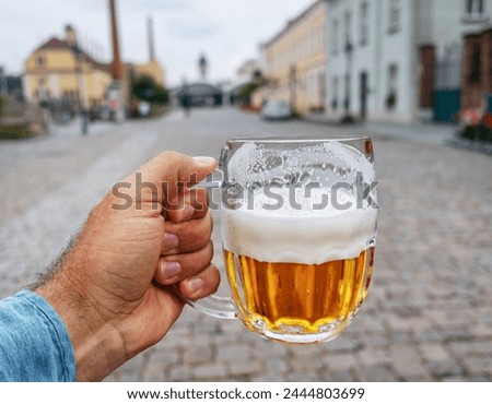 A man holds  mug of beer in his hand and blurred paving stones and old-fashioned houses at the background.