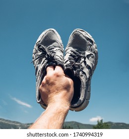 Man Holds Old Running Sneakers In Hand On Background Of BBue Sky And Mountains