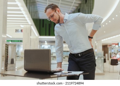 A Man Holds On To His Back, Suffers From Lower Back Pain, Standing Near A Desktop With A Computer. Sedentary Work Office Worker