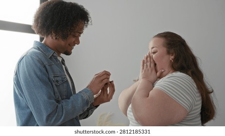Man holds open ring box and slides ring onto woman's hand while she clasps hands to chest in building; joy love commitment.