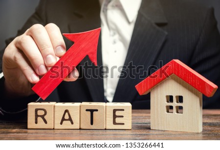 A man holds a red arrow up above the word Rate and a wooden house. The concept of raising interest rates on mortgages. The increase in property tax rates. Real estate capitalization. Insurance.