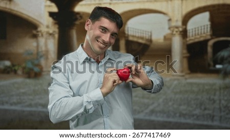 Man holds red heart between fingers in a sunlit building courtyard with arched columns, smiling gently; romance.