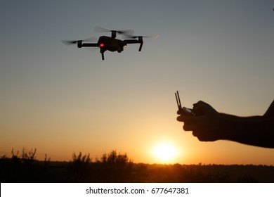 Man holds remote controller with his hands while copter is flying on background. Drone hovers behind the pilot on suset. No face