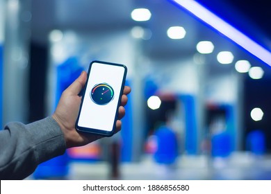 A Man Holds A Smartphone With A Digital Fuel Meter On The Screen Against The Background Of A Night Gas Station For A Car