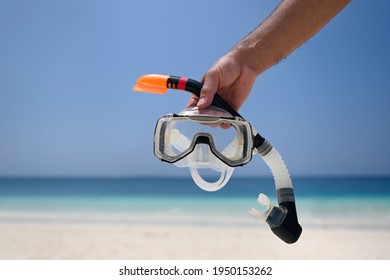 A man holds a snorkel mask in his hand against a clear blue sky and turquoise ocean. Diving mask in a man's hand. Photo of a mask and snorkel for swimming in a pool or sea with a central composition