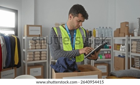 Man holds tablet when checking items in center where hispanic young volunteer catalogs donations and guy arranges clothes for local charity.