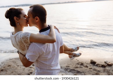 A Man Holds A Woman In His Arms And Kisses. A Young Girl Hugs The Man Tenderly By The Neck. A Loving Couple Kissing. People Stand By The River. High Quality Photo