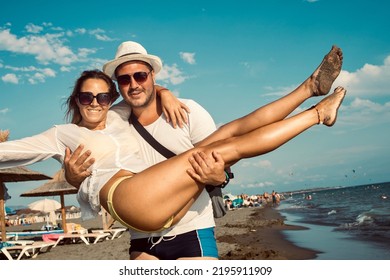 A Man Holds A Woman In His Arms On The Beach. Both The Man And The Woman Wear Sunglasses And The Man Has A Hat. They Both Smile. They Enjoy Their Vacation