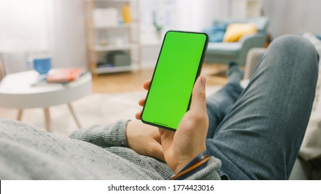 Man At Home Lying On A Couch Using Smartphone With Green Mock-up Screen. Over The Shoulder Camera Shot.