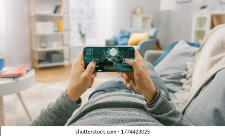 Man at Home Lying on a Couch using Smartphone, Holds it Horizontally in Landscape Mode. He is Playing First Person Shooter Video Game. Point of View Camera Shot.
