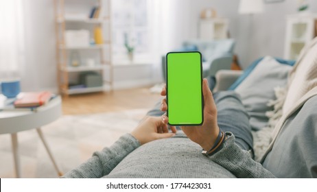 Man At Home Lying On A Couch Using Smartphone With Green Mock-up Screen, Doing Swiping, Scrolling Gestures. Guy Using Mobile Phone, Internet Social Networks Browsing. Point Of View Shot.