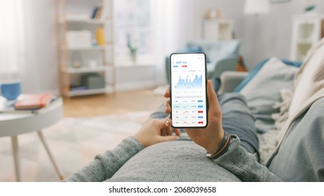 Man At Home Lying On A Couch And Using Smartphone With Stock Market App To Check Share Prices. Graph Is Going Up Making A Great Profit. In The Background Cozy Homely Atmosphere.