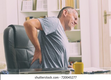 Man In Home Office Suffering From Low Back Pain Sitting At The Desk With Notebook, Papers And Other Objects