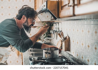 Man at home tasting food from the pot with a table spoon. People in the kitchen preparing dinner. One mature male alone cooking. Real lifestyle person testing taste and cook preparation. Husband work - Powered by Shutterstock