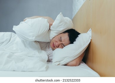 A Man At Home Tries To Fall Asleep, Noisy Neighbors Interfere With Sleep, An Asian Closes His Ears With Pillows, Tired After Work