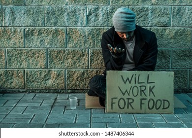 A Man, Homeless, A Man Asks For Alms On The Street With A Sign Will Work For Food. Concept Of Homeless Person, Addict, Poverty, Despair