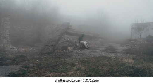 Man in a hood sitting among ruins at mystical mist, post-apocalyptic scene