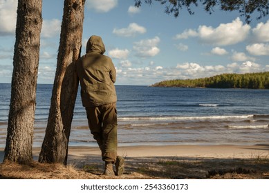 Man in Hooded Forest Attire by Tranquil Lakeside. A back view of a person clad in a hooded forest outfit, standing on a sandy lakeshore. The traveler enjoys a beautiful view of the lake - Powered by Shutterstock