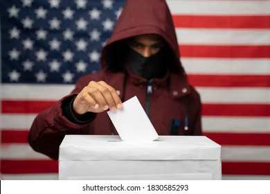 Man In Hoodie With Face Covered Casting Vote At Polling Booth With US Falg As Background - Concept Of Unknown Voting Or Vote Rigging In US Elections.