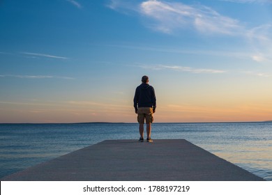 Man in hoodie and short cargo pants standing on dock and looking on sea horizon, island od Pag, Adriatic sea, Croatia
