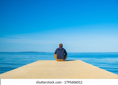 Man in hoodie and short cargo sitting on the edge of dock and meditating. Sea horizon on the island od Pag, Adriatic sea, Croatia. Early morning meditation.
