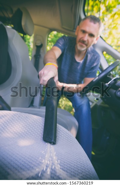 Man hoovering a seat inside a car\
cabin, visible dirt. Cleaning car with vacuum\
cleaner.