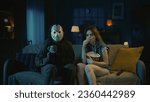 Man horror maniac in white mask and girl with popcorn sitting on the sofa watching movie in the dark apartment room.