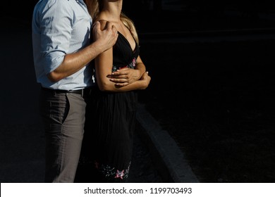 Man Hugs Girl From Behind On The Street, Hugging Her, Attracted To Each Other. Girl In A Black Dress. Man Hugs From Behind The Woman And Kisses On The Forehead.the Couple Is Standing Against A Dark