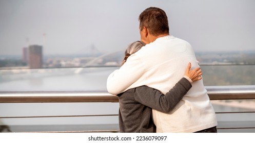 A man hugs a woman, an elderly couple happily spend time together on a walk, embracing look at the city landscape through the railing of a modern bridge, husband and wife support each other. - Shutterstock ID 2236079097