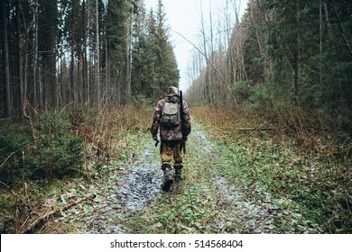 Man the hunter goes through the forest in rubber boots, with a backpack and a gun. Cloudy weather, autumn. On the ground, a little of snow. View from the back.